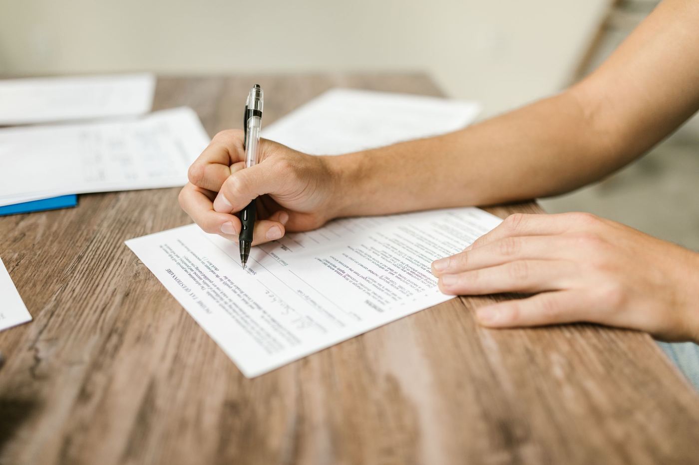 A hand filling in a paper form with a pen at a wooden desk